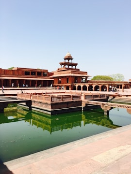 A historic architectural structure made of red sandstone set against a clear blue sky. The building features intricately carved pillars and archways. A rectangular pool with greenish water reflects the structure. Visitors can be seen walking around the complex.