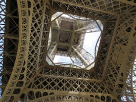 A symmetrical view of an intricate lattice structure made of iron, looking up towards the sky. The geometric patterns and detailed metalwork create a complex and industrial aesthetic. Light filters through the gaps, adding contrast and depth to the design.