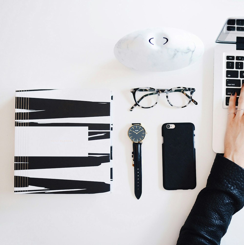 A flatlay image of a white desk with a laptop, reading glasses, a watch, smartphone and various other items.