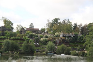 Lush green vegetation covers a landscape with multiple terraced levels, surrounded by trees. A body of water is visible in the foreground, reflecting the greenery. Stone structures and pathways are integrated into the scene, blending with the natural environment.