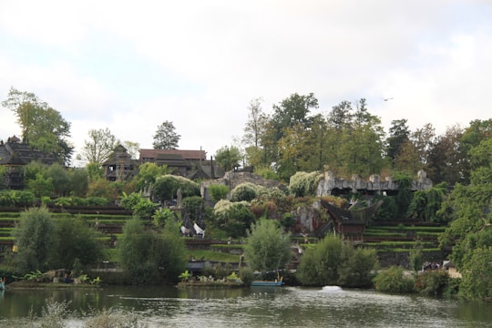 Lush green vegetation covers a landscape with multiple terraced levels, surrounded by trees. A body of water is visible in the foreground, reflecting the greenery. Stone structures and pathways are integrated into the scene, blending with the natural environment.