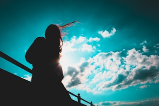 A silhouette of a person stands against a bright sky filled with clouds. The person's hair appears to be blowing in the wind, and they are leaning against a railing.