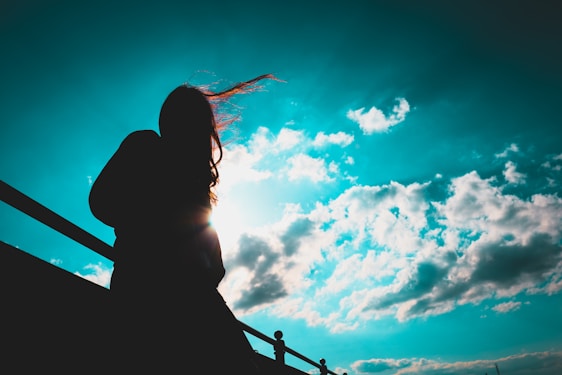 A silhouette of a person stands against a bright sky filled with clouds. The person's hair appears to be blowing in the wind, and they are leaning against a railing.