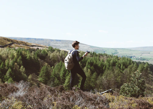 A warm photo of a traveler holding a camera, smiling against a backdrop of lush green hills.