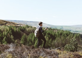 A person stands on a hill with a camera in hand, surrounded by lush greenery and an expansive view of rolling hills and forests. The individual carries a backpack, suggesting they are on a hiking or photography excursion. The scene conveys a sense of adventure and tranquility.