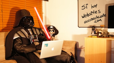A person in a black costume resembling Darth Vader is sitting on a couch using a laptop. They are holding a red lightsaber in their left hand. There is a whiteboard in the background with handwritten text in Spanish that reads 'Sí, hay websites acroachcode.com'. A plush toy dressed as a pirate and a raccoon toy are on a small wooden cabinet next to a telephone and some papers.