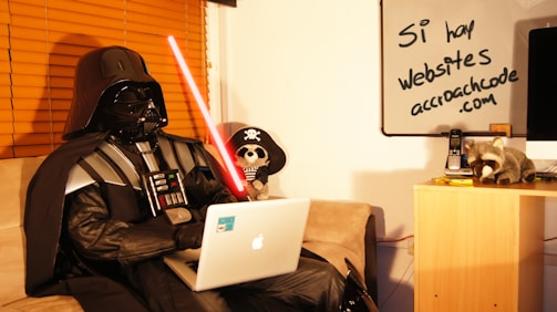 A person in a black costume resembling Darth Vader is sitting on a couch using a laptop. They are holding a red lightsaber in their left hand. There is a whiteboard in the background with handwritten text in Spanish that reads 'Sí, hay websites acroachcode.com'. A plush toy dressed as a pirate and a raccoon toy are on a small wooden cabinet next to a telephone and some papers.