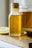 A glass bottle with a cork lid contains a golden liquid labeled as 'Four Ounces Pure Honey'. The bottle is placed on a wooden surface next to a sliced lemon, a jar of the same golden liquid, and a metal measuring spoon with a trace of the liquid on its surface.