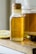 A glass bottle with a cork lid contains a golden liquid labeled as 'Four Ounces Pure Honey'. The bottle is placed on a wooden surface next to a sliced lemon, a jar of the same golden liquid, and a metal measuring spoon with a trace of the liquid on its surface.