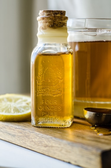 A glass bottle with a cork lid contains a golden liquid labeled as 'Four Ounces Pure Honey'. The bottle is placed on a wooden surface next to a sliced lemon, a jar of the same golden liquid, and a metal measuring spoon with a trace of the liquid on its surface.