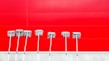 Seven mops with metal handles are lined up against a vibrant red wall. The mops are neatly arranged, casting subtle shadows on the wall, creating a pattern of lines and textures.