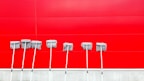 Seven mops with metal handles are lined up against a vibrant red wall. The mops are neatly arranged, casting subtle shadows on the wall, creating a pattern of lines and textures.