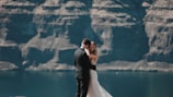 A bride and groom embrace against a backdrop of towering rocky cliffs and a calm body of water. The bride wears a white gown, and the groom is in a dark suit. The scene conveys a sense of intimacy and natural beauty.