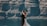 A bride and groom embrace against a backdrop of towering rocky cliffs and a calm body of water. The bride wears a white gown, and the groom is in a dark suit. The scene conveys a sense of intimacy and natural beauty.