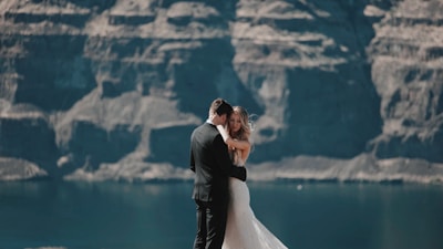 A bride and groom embrace against a backdrop of towering rocky cliffs and a calm body of water. The bride wears a white gown, and the groom is in a dark suit. The scene conveys a sense of intimacy and natural beauty.