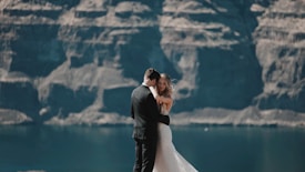 A bride and groom embrace against a backdrop of towering rocky cliffs and a calm body of water. The bride wears a white gown, and the groom is in a dark suit. The scene conveys a sense of intimacy and natural beauty.