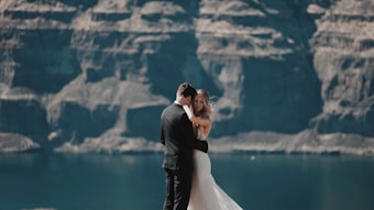A bride and groom embrace against a backdrop of towering rocky cliffs and a calm body of water. The bride wears a white gown, and the groom is in a dark suit. The scene conveys a sense of intimacy and natural beauty.