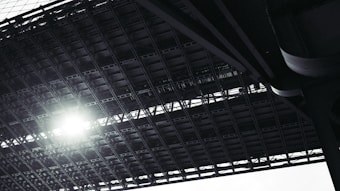 A close-up view from below a large steel structure, likely a bridge, with beams and girders crossing overhead. Sunlight filters through, creating a bright spot among the dark metal elements.