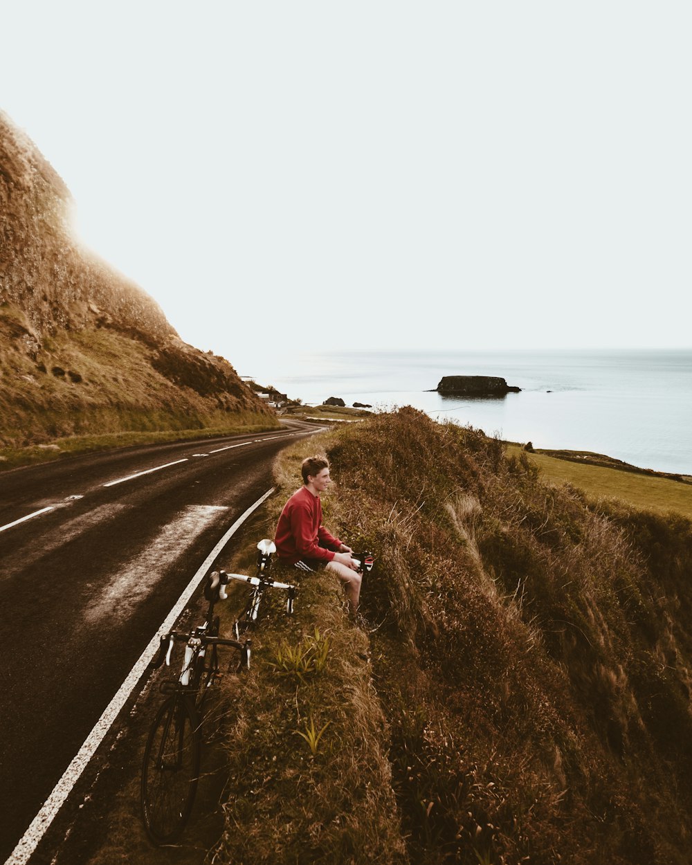 woman in red jacket and white pants sitting on gray concrete road near body of water