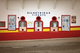 A building facade with ticket windows and vibrant color schemes. The wall features three arched windows with glass blocks and showcases old posters and advertisements around them. The colors are primarily white, red, and yellow. Above the windows, the words 'Bilheteiras' and 'Tickets' are displayed in blue lettering.