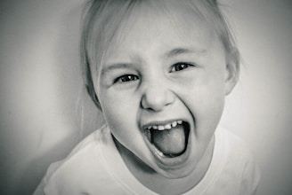 A black and white close-up image of a young child with blonde hair. The child is displaying a wide, open-mouthed expression that conveys excitement or joy, with eyes open wide. The background is a soft, out-of-focus white, enhancing the focus on the child's face.