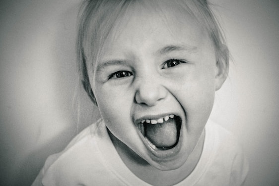 A black and white close-up image of a young child with blonde hair. The child is displaying a wide, open-mouthed expression that conveys excitement or joy, with eyes open wide. The background is a soft, out-of-focus white, enhancing the focus on the child's face.
