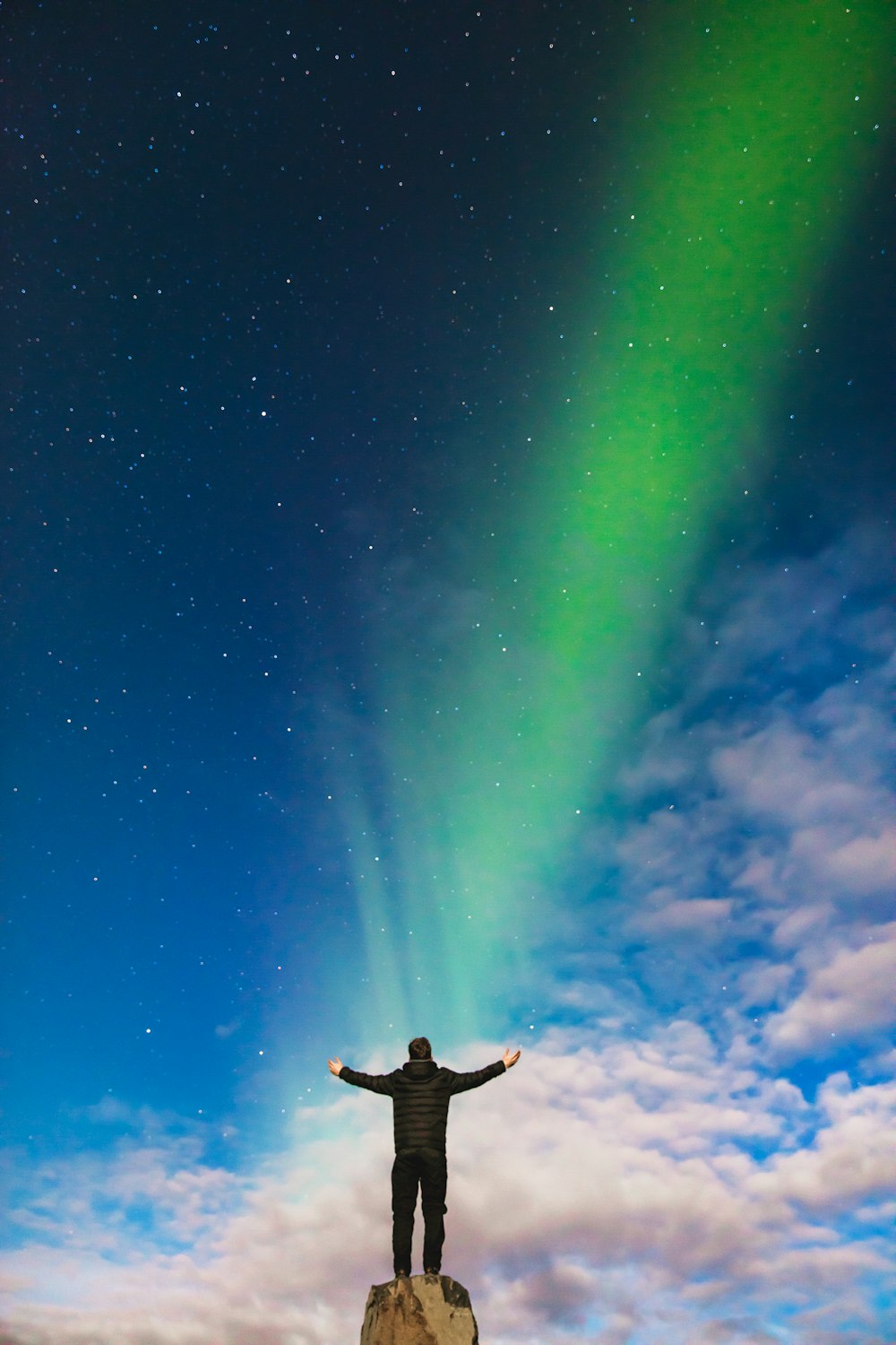 Mann steht auf Felsen unter grüner Aurora Borealis und weißen Wolken