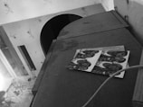 A black and white image shows a cluttered indoor scene with a table that has various items including papers and a bottle. A person is sitting on the left side holding a newspaper or book. On top of a cabinet, there is a piece of paper with multiple identical photos of people.
