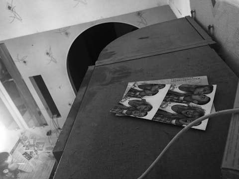 A black and white image shows a cluttered indoor scene with a table that has various items including papers and a bottle. A person is sitting on the left side holding a newspaper or book. On top of a cabinet, there is a piece of paper with multiple identical photos of people.