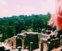 Ancient ruins with stone arches and walls are surrounded by dense forest. A noticeable tuft of pink hair appears on the right side of the image against a backdrop of greenery and scattered clouds.