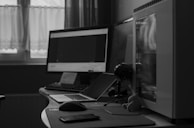 A monochrome photograph of a computer setup on a desk. The setup includes two monitors, a desktop computer chassis with a transparent side panel, a laptop, a keyboard, a mouse, and a smartphone. A small plush toy and a microphone stand are visible next to the computer. The background features a curtained window allowing natural light into the room.