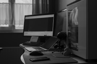 A monochrome photograph of a computer setup on a desk. The setup includes two monitors, a desktop computer chassis with a transparent side panel, a laptop, a keyboard, a mouse, and a smartphone. A small plush toy and a microphone stand are visible next to the computer. The background features a curtained window allowing natural light into the room.