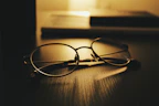 A pair of round eyeglasses resting on a dark wooden surface, illuminated by warm, soft lighting. In the background, there are out-of-focus books or notebooks, creating a cozy and contemplative atmosphere.