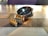 A silver wristwatch with a shiny metal bracelet lies on a wooden table with a checkered pattern. The watch face is dark with a subtle design and the brand name 'Daniel Klein' is visible. The lighting creates a warm reflection on the watch.