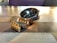 A silver wristwatch with a shiny metal bracelet lies on a wooden table with a checkered pattern. The watch face is dark with a subtle design and the brand name 'Daniel Klein' is visible. The lighting creates a warm reflection on the watch.