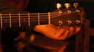 A close-up view of a hand holding the headstock of an acoustic guitar with metallic tuning pegs. The background is dimly lit, creating a warm, intimate ambiance.