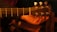 A close-up view of a hand holding the headstock of an acoustic guitar with metallic tuning pegs. The background is dimly lit, creating a warm, intimate ambiance.
