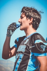 A young football player wearing a black and blue jersey with the number 77, holding a blue mouthguard near his mouth. His hair is slightly messy and he is wearing one glove. The background features a clear blue sky and distant hills.