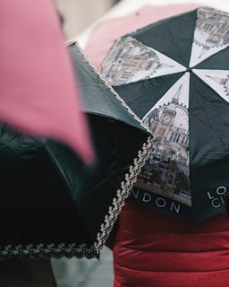 Two umbrellas are prominently visible, one featuring illustrations of iconic London landmarks including Big Ben and the London Eye, and the other with a decorative lace trim. Both umbrellas are wet, indicating rainfall, and the lower portion of a person is visible wearing a red coat.