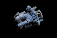 An astronaut wearing a white spacesuit floats in space, using a jetpack for maneuvering. The reflective golden visor of the helmet contrasts against the dark backdrop of space.