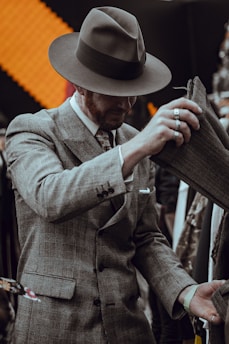A man dressed in a vintage-style gray plaid suit and a wide-brimmed hat is examining fabric. He wears multiple silver rings and a patterned tie. The background features an orange textured pattern and various hanging clothes.
