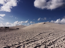 Sand dunes stretch across the horizon under a partly cloudy sky. Several vehicles are visible on the sandy terrain, suggesting an off-road adventure in a desert landscape.
