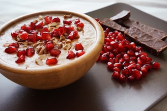 A wooden bowl filled with a creamy dessert topped with pomegranate seeds and nuts sits on a dark surface. Beside the bowl, there are several pieces of chocolate and a pile of loose pomegranate seeds, adding a rich contrast of textures and colors.