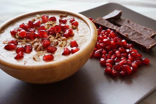 A wooden bowl filled with a creamy dessert topped with pomegranate seeds and nuts sits on a dark surface. Beside the bowl, there are several pieces of chocolate and a pile of loose pomegranate seeds, adding a rich contrast of textures and colors.
