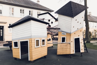 A playground structure with unique, tilted towers made of wood and painted in white and beige. The design includes small windows and climbing holds, set in an outdoor area next to residential buildings.