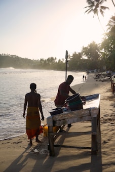 Two people are near the shore, with one person working at a table and another standing close by. The beach is lined with palm trees, and there are a few people in the distance walking or relaxing. The ocean waves are gently crashing onto the sandy beach, and the setting sun casts a warm glow over the scene.