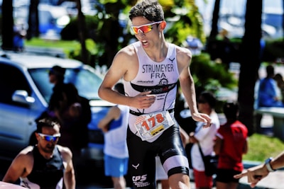 A triathlete is captured in motion during a race, wearing sport sunglasses and athletic gear with a race number. Surrounding the main athlete are blurred figures of spectators and a parked vehicle, suggesting a lively event atmosphere.