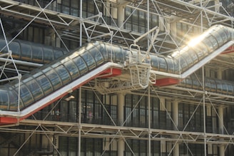 A modern architectural structure features a prominent glass and steel tube on the outside, angled diagonally and supported by a network of exposed metallic beams. Sunlight reflects off the glass surface, creating a bright spot on the transparent tube. In the background, grid-like windows and further structural elements are visible.