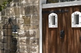 A rustic wooden door with two small, arched glass windows framed in white and decorated with iron embellishments. Adjacent to the door is a stone wall displaying a carved stone detail and a black wrought iron grille. The overall setting suggests an entrance with a charming, historic appearance.