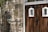 A rustic wooden door with two small, arched glass windows framed in white and decorated with iron embellishments. Adjacent to the door is a stone wall displaying a carved stone detail and a black wrought iron grille. The overall setting suggests an entrance with a charming, historic appearance.